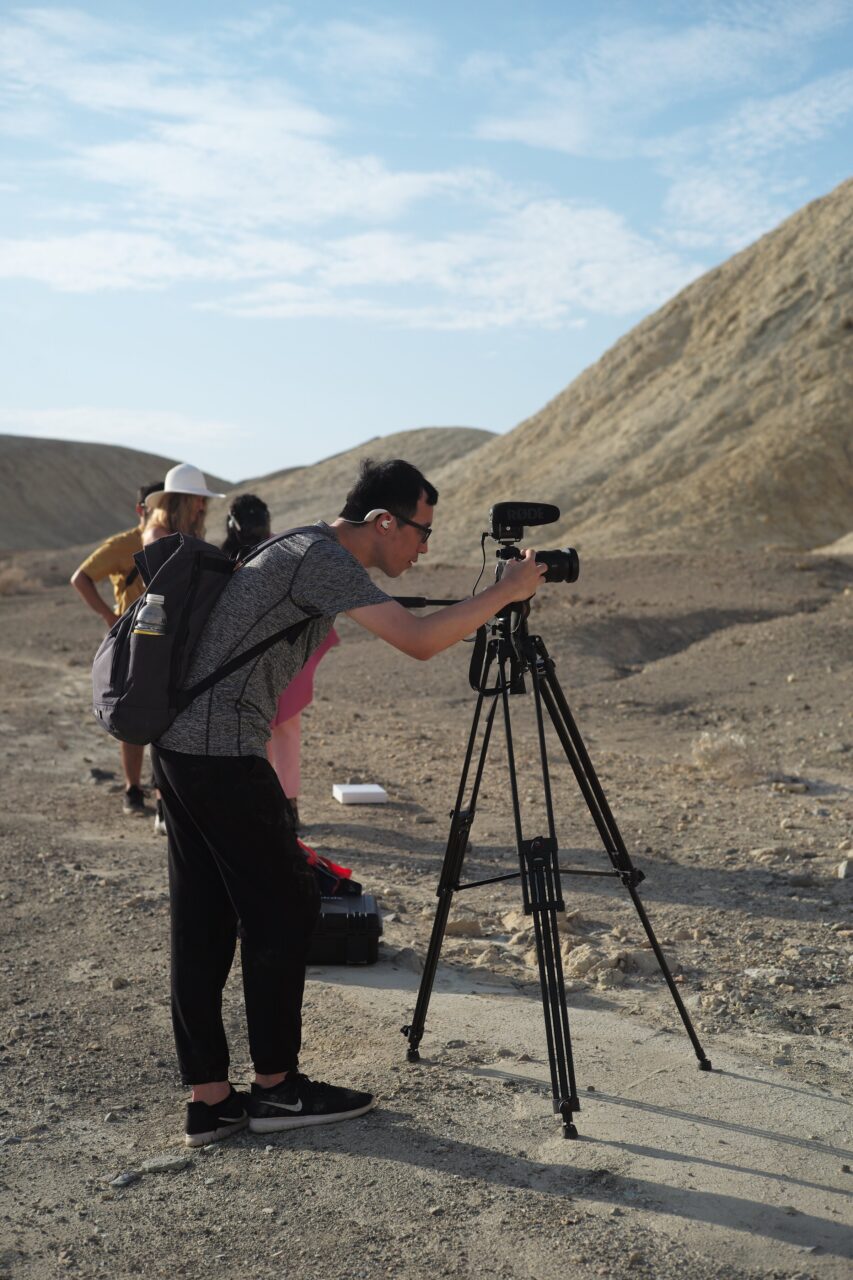 Videographer adjusting a camera on a tripod in front of pale desert hills