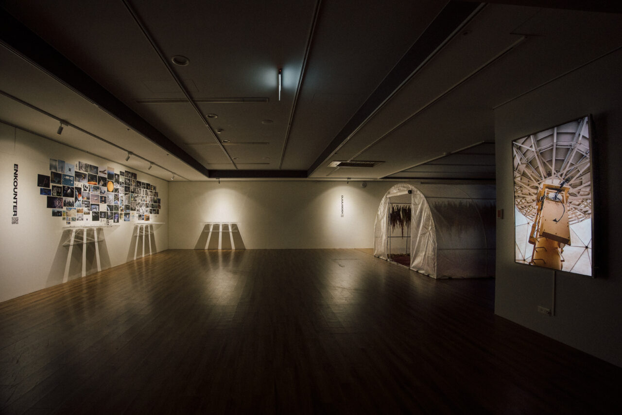 Installation view with the ENKOUNTER document wall, the djulis greenhouse, and a screen showing the satellite antenna filmed at Taiwan's National Space Organization