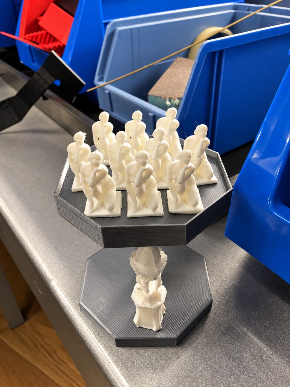 Rows of small white cast figures on a black octagonal stand, with a single figure under the stand, beside blue bins.