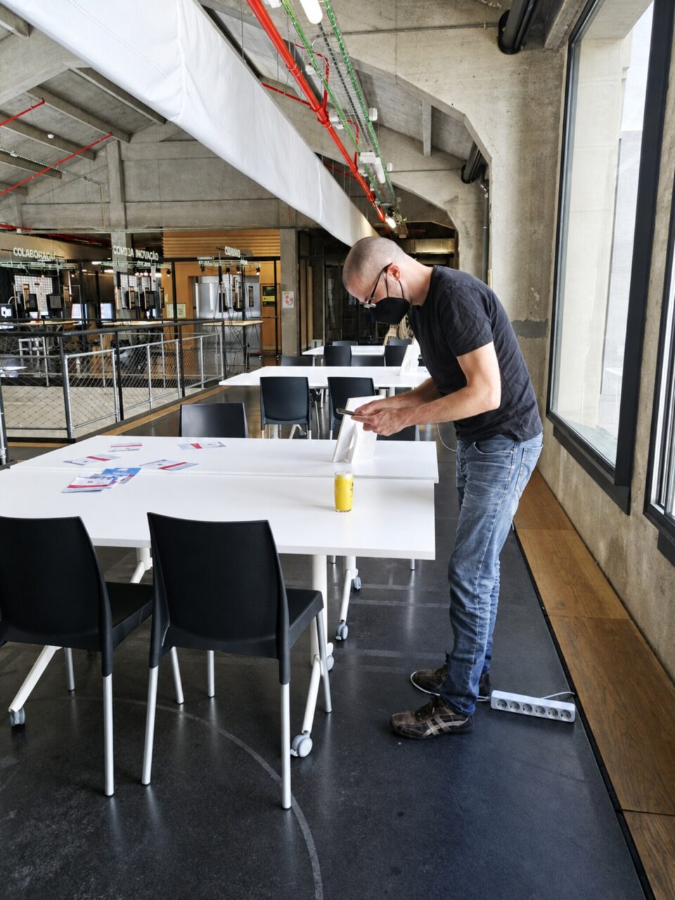 A man in a mask bends over a white table photographing a small object with a phone, in a large hall.