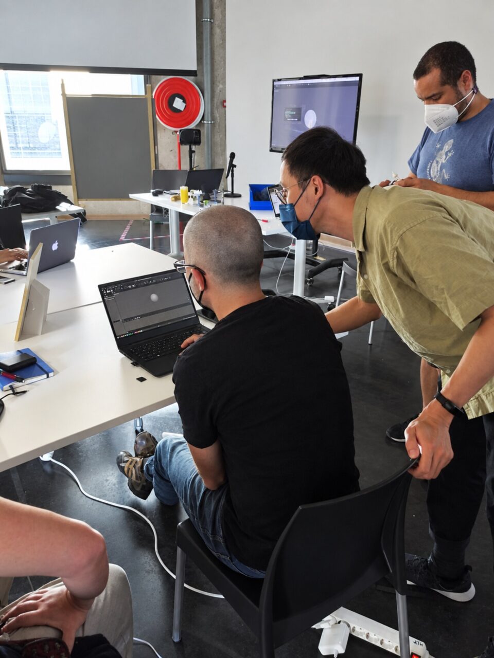 A masked facilitator leans over a seated participant at a laptop while another participant watches.