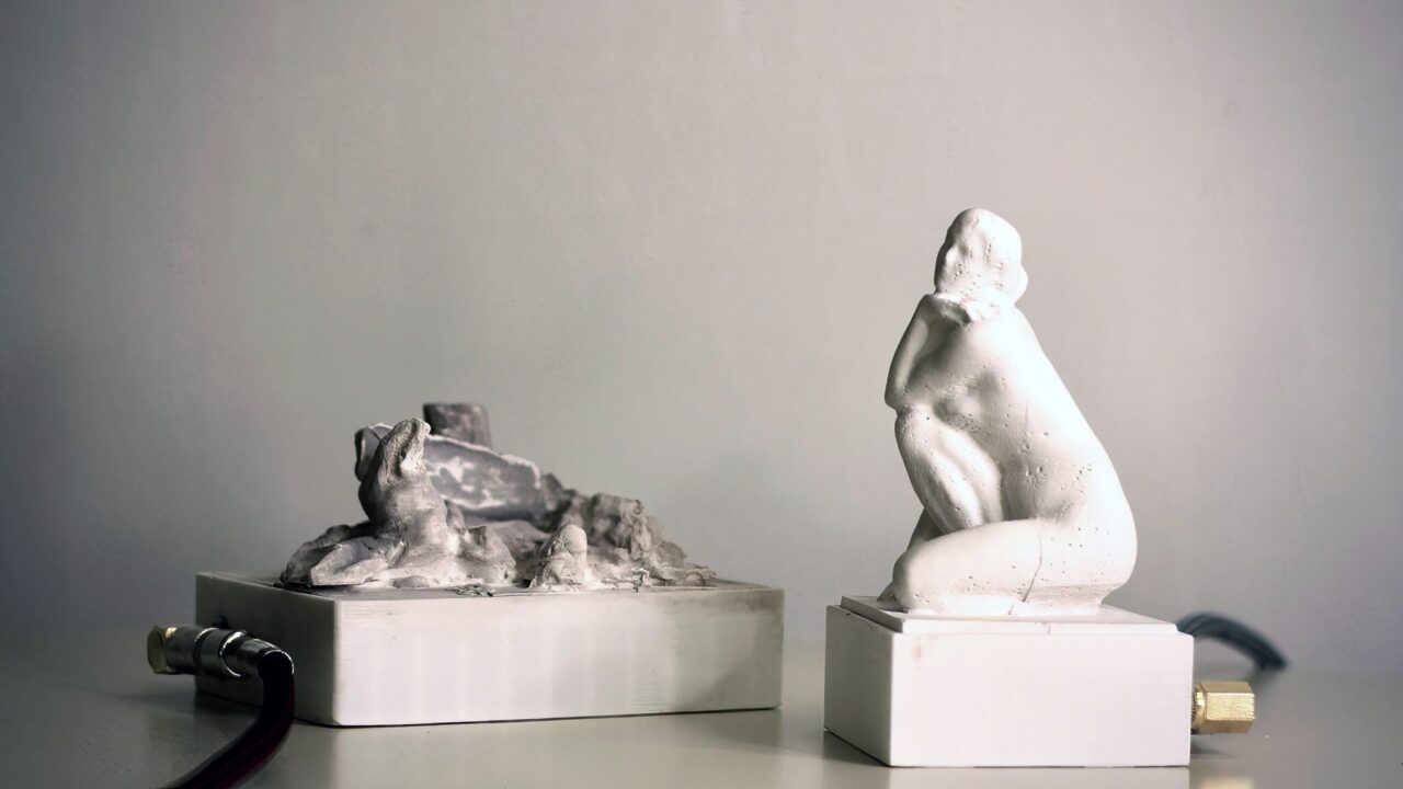 Two white cast objects on plinths, a rocky landscape form and a kneeling figure, with cables and brass connectors.