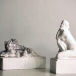 Two white cast objects on plinths, a rocky landscape form and a kneeling figure, with cables and brass connectors.