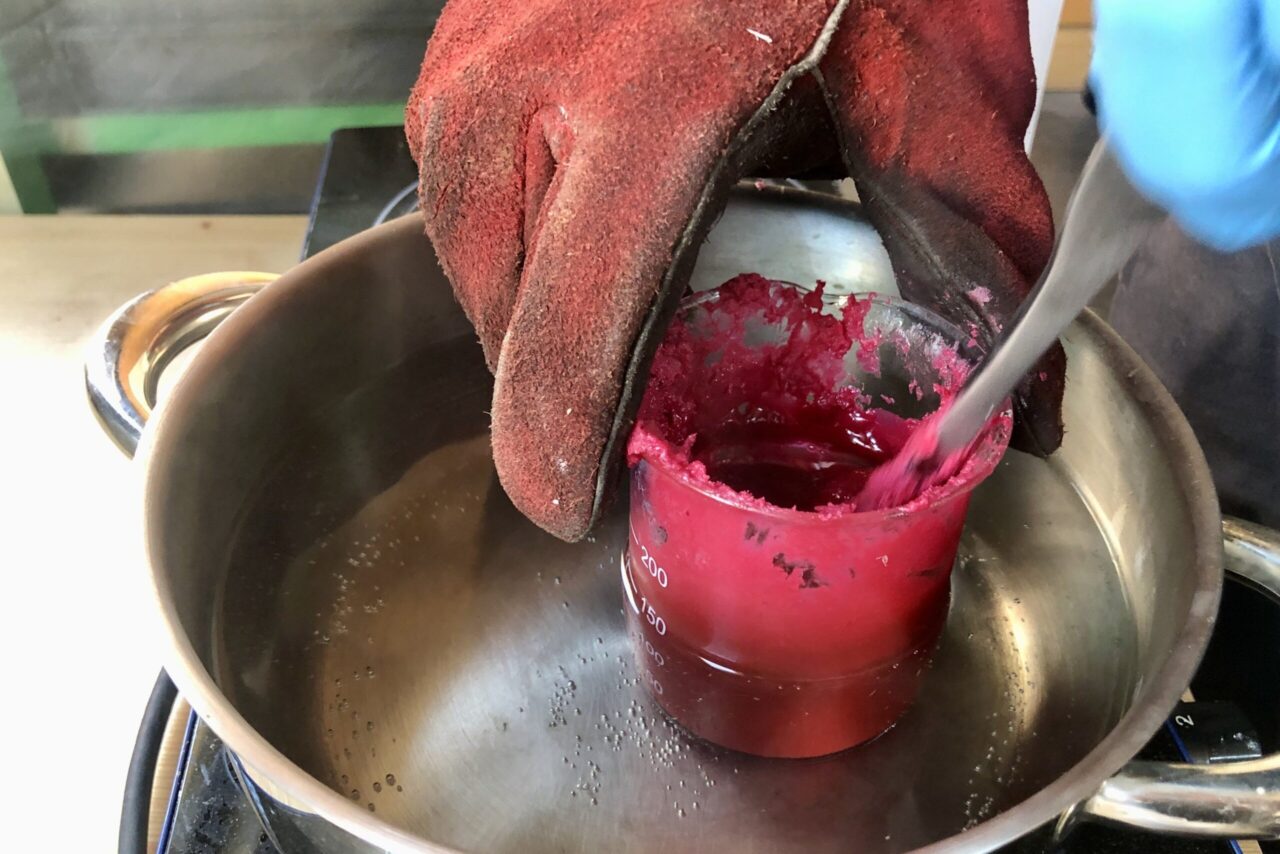 Gloved hand stirring melted red wax in a beaker set in a pot of water on a hotplate.