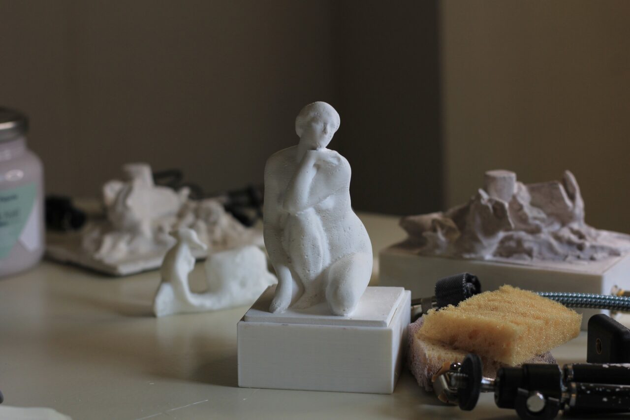 White kneeling figure on a square plinth on a table, with other cast pieces, a sponge and cables blurred around it.