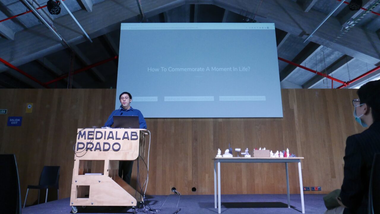 Speaker at a Medialab Prado podium on a dark stage, a large projection screen behind and cast objects on a side table.