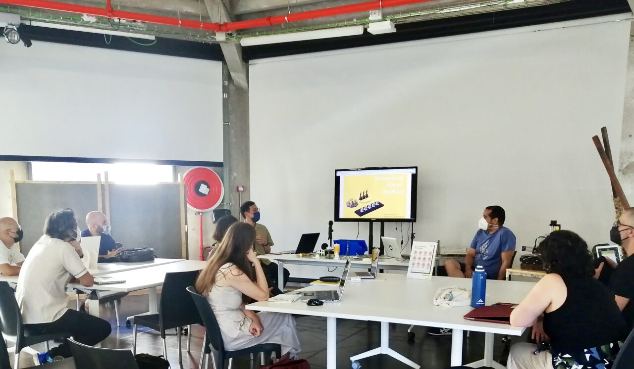 Workshop room with participants seated at white tables facing a screen showing 3D models, windows behind.