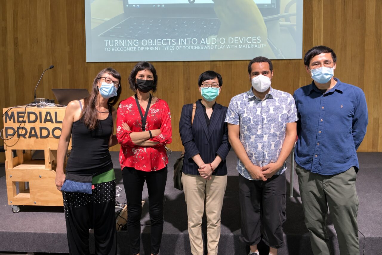 Five masked attendees posing together beside the Medialab Prado lectern, with a slide about objects and audio interfaces projected behind them.